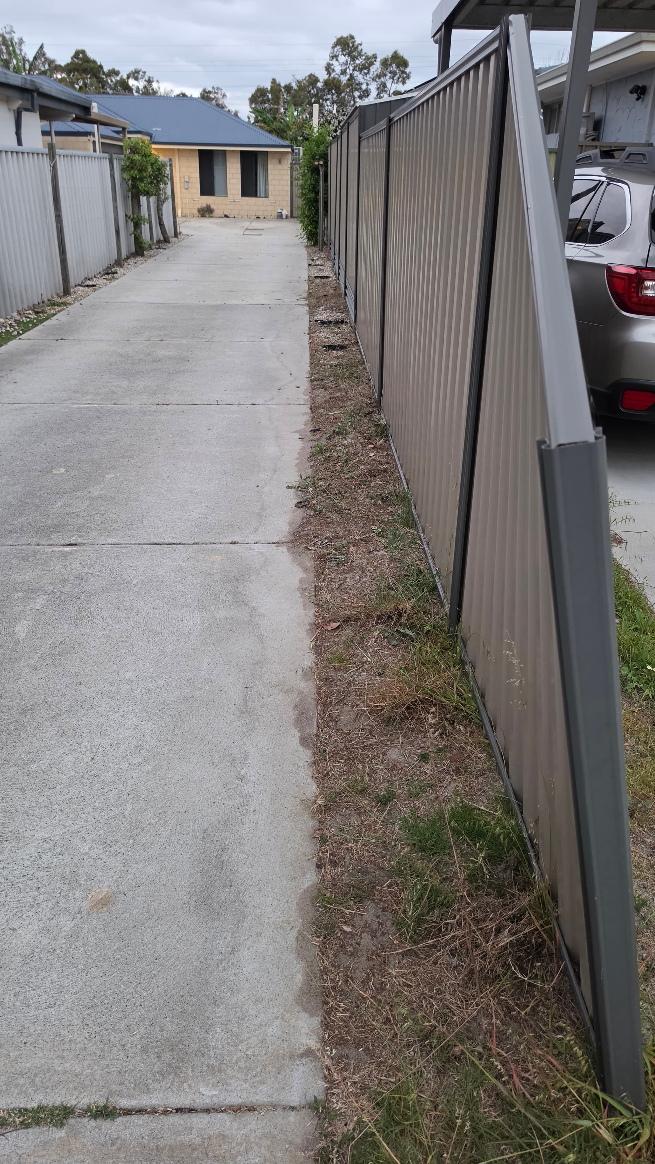 Long concrete driveway beside a corrugated metal fence with a leaning grey post cover.